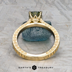 The "Maya" ring in 18k yellow gold features ornate engravings and a 1.48-carat Australian sapphire, shown upright on a textured surface with a blue stone in the background.