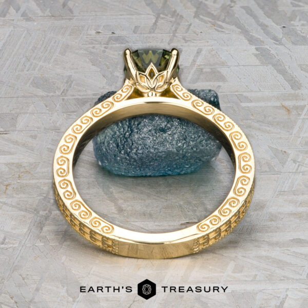 The "Maya" ring in 18k yellow gold with 1.48-carat Australian sapphire The "Maya" ring in 18k yellow gold features ornate engravings and a 1.48-carat Australian sapphire, shown upright on a textured surface with a blue stone in the background.