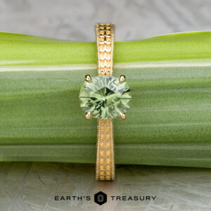 The "Maya" ring in 18k yellow gold with a 1.48-carat Australian sapphire stands upright against a green leaf background. The text "EARTH'S TREASURY" is displayed at the bottom.