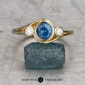 The "Maeve" ring in 14k yellow gold, featuring a 0.91-carat Montana sapphire center and two white accent stones, is displayed upright on a rough blue mineral with the "Earth's Treasury" logo below.