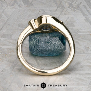 The "Maeve" ring, crafted in 14k yellow gold with a 0.91-carat uncut Montana sapphire, stands upright on a textured gray surface. "EARTH'S TREASURY" is displayed at the bottom of the image.