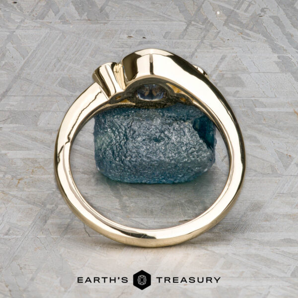 The "Maeve" ring in 14k yellow gold with 0.91-carat Montana sapphire The "Maeve" ring, crafted in 14k yellow gold with a 0.91-carat uncut Montana sapphire, stands upright on a textured gray surface. "EARTH'S TREASURY" is displayed at the bottom of the image.