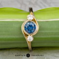 The "Maeve" ring in 14k yellow gold features a 0.91-carat Montana sapphire at the center with two smaller white stones on either side, shown against a green leaf background.