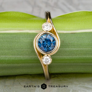 The "Maeve" ring in 14k yellow gold features a 0.91-carat Montana sapphire at the center with two smaller white stones on either side, shown against a green leaf background.