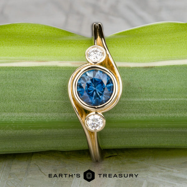 The "Maeve" ring in 14k yellow gold with 0.91-carat Montana sapphire The "Maeve" ring in 14k yellow gold features a 0.91-carat Montana sapphire at the center with two smaller white stones on either side, shown against a green leaf background.