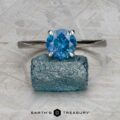 The "Elena" ring in 14k white gold with a 1.93-carat Montana sapphire is displayed above a rough blue crystal on a gray textured surface, with the "Earth's Treasury" logo below.