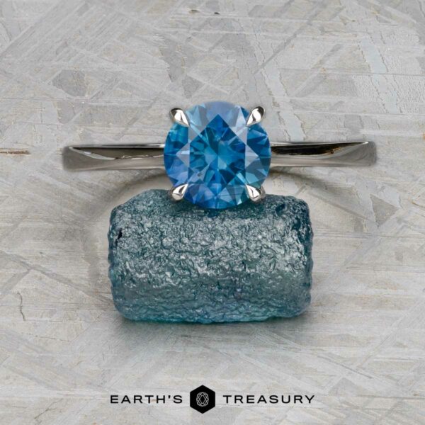 The "Elena" ring in 14k white gold with 1.93-carat Montana sapphire The "Elena" ring in 14k white gold with a 1.93-carat Montana sapphire is displayed above a rough blue crystal on a gray textured surface, with the "Earth's Treasury" logo below.