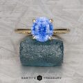 A gold cathedral solitaire ring by Benyo, Scott and McIsaac, Darcy with a blue oval-cut gemstone rests on a rough green stone, set against a textured light grey background. "Earth's Treasury" appears at the bottom.