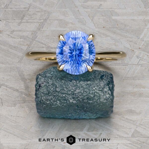 Benyo, Scott and McIsaac, Darcy A gold cathedral solitaire ring by Benyo, Scott and McIsaac, Darcy with a blue oval-cut gemstone rests on a rough green stone, set against a textured light grey background. "Earth's Treasury" appears at the bottom.