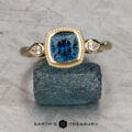 The "Sadie" ring in 14k yellow gold, featuring a 1.22-carat Montana sapphire and two round side diamonds, is displayed atop a rough blue stone with "Earth's Treasury" branding below—a refined three-stone ring design.