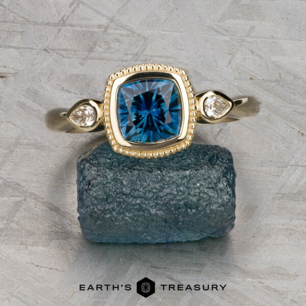 The "Sadie" ring in 14k yellow gold with 1.22-carat Montana sapphire The "Sadie" ring in 14k yellow gold, featuring a 1.22-carat Montana sapphire and two round side diamonds, is displayed atop a rough blue stone with "Earth's Treasury" branding below—a refined three-stone ring design.