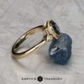 The "Sadie" ring in 14k yellow gold with a 1.22-carat Montana sapphire is displayed beside a rough blue stone on a textured surface, with the Earth’s Treasury logo visible at the bottom.
