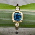 The "Sadie" ring in 14k yellow gold showcases a 1.22-carat Montana sapphire center, flanked by two pear-shaped clear stones, beautifully displayed on a green leaf.