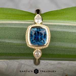 The "Sadie" ring in 14k yellow gold showcases a 1.22-carat Montana sapphire center, flanked by two pear-shaped clear stones, beautifully displayed on a green leaf.