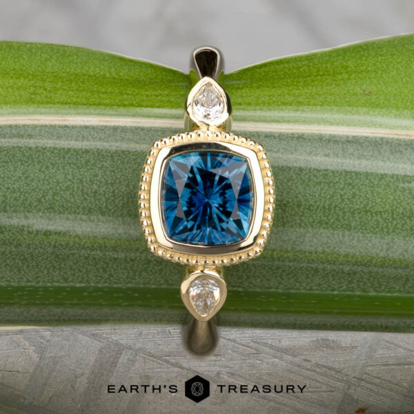 The "Sadie" ring in 14k yellow gold with 1.22-carat Montana sapphire The "Sadie" ring in 14k yellow gold showcases a 1.22-carat Montana sapphire center, flanked by two pear-shaped clear stones, beautifully displayed on a green leaf.