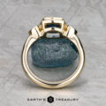 The "Sadie" ring in 14k yellow gold with a 1.22-carat Montana sapphire stands upright against a textured stone backdrop on a gray patterned surface. "EARTH'S TREASURY" is displayed at the bottom.