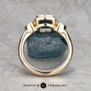 The "Sadie" ring in 14k yellow gold with a 1.22-carat Montana sapphire stands upright against a textured stone backdrop on a gray patterned surface. "EARTH'S TREASURY" is displayed at the bottom.