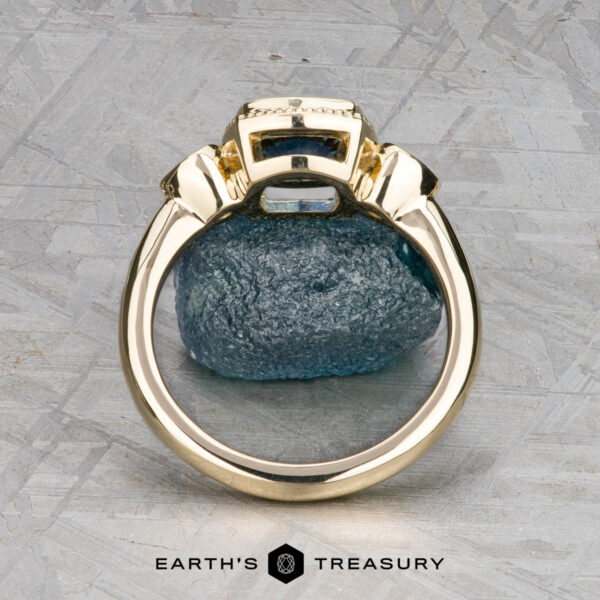 The "Sadie" ring in 14k yellow gold with 1.22-carat Montana sapphire The "Sadie" ring in 14k yellow gold with a 1.22-carat Montana sapphire stands upright against a textured stone backdrop on a gray patterned surface. "EARTH'S TREASURY" is displayed at the bottom.