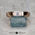 The "Theo" ring in 14k rose gold with a 0.48-carat heirloom diamond is balanced on a raw blue-green gemstone against a textured gray background, with the "Earth's Treasury" logo at the bottom.