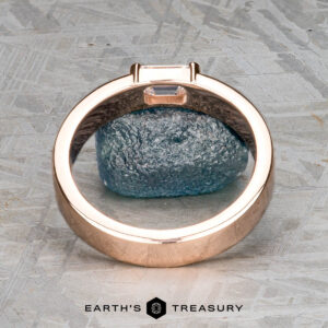 The "Theo" ring in 14k rose gold features a 0.48-carat heirloom diamond set upright and is photographed on a textured gray surface with a rough blue stone in the background.