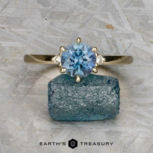 The "Samia" ring in 14k yellow gold with 1.73-carat Montana sapphire The "Samia" ring in 14k yellow gold, featuring a 1.73-carat Montana sapphire and two small side diamonds, is displayed on a rough blue stone above the Earth's Treasury logo.
