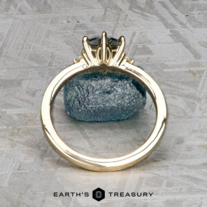 The "Samia" ring in 14k yellow gold, featuring a 1.73-carat Montana sapphire, is displayed vertically on a blue stone surface with a geometric patterned background and the Earth's Treasury logo below.