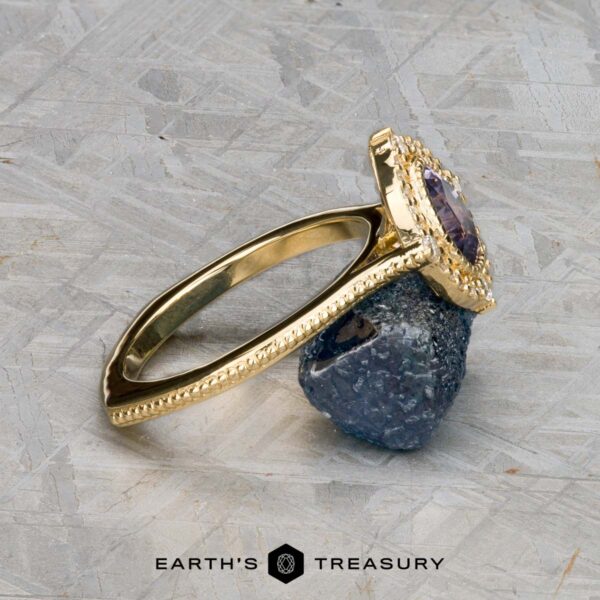 The "Marietta" ring in 18k yellow gold with 1.13-carat Montana sapphire The "Marietta" ring in 18k yellow gold, featuring a 1.13-carat rectangular Montana sapphire, rests on a rough dark stone atop a textured gray surface. The Earth's Treasury logo is shown at the bottom.