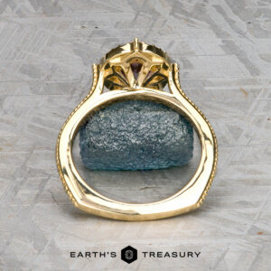 The "Marietta" ring in 18k yellow gold with a 1.13-carat Montana sapphire stands upright on a rough blue stone surface, with Earth's Treasury logo at the bottom.