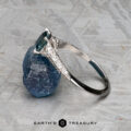 The "Anya" ring in 14k white gold with a 0.74-carat Montana sapphire stands upright, leaning against a rough blue stone on a gray textured surface. The Earth's Treasury logo is featured at the bottom.