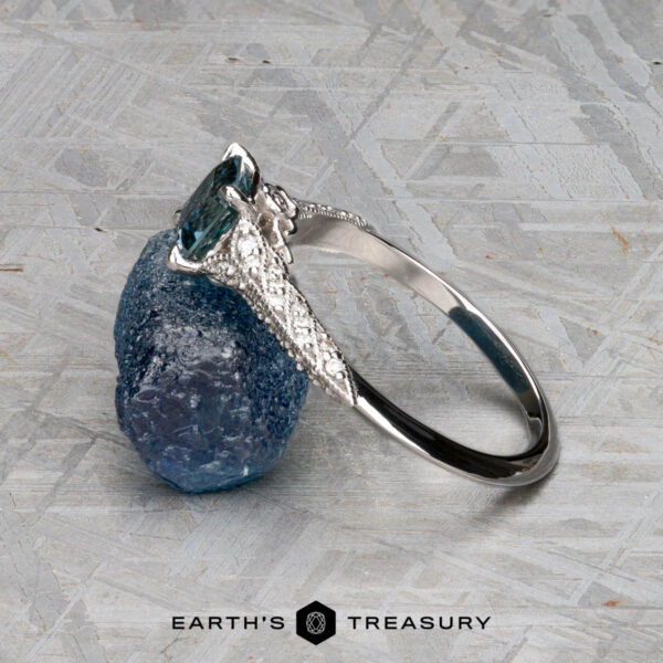 The "Anya" ring in 14k white gold with 0.74-carat Montana sapphire The "Anya" ring in 14k white gold with a 0.74-carat Montana sapphire stands upright, leaning against a rough blue stone on a gray textured surface. The Earth's Treasury logo is featured at the bottom.