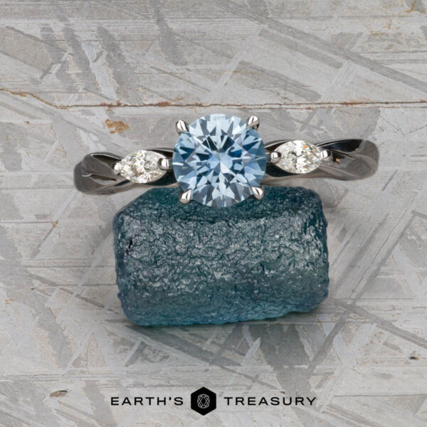 The Braided "Zuri" ring in 14k white gold with 1.15-carat Montana sapphire The Braided "Zuri" ring in 14k white gold, featuring a 1.15-carat Montana sapphire, is displayed on a rough blue crystal atop a textured gray surface.