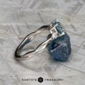 The Braided "Zuri" ring in 14k white gold with a 1.15-carat Montana sapphire is displayed against a rough blue stone on a light textured surface. The logo reads "Earth's Treasury.