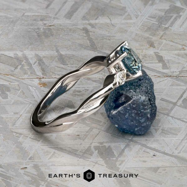 The Braided "Zuri" ring in 14k white gold with 1.15-carat Montana sapphire The Braided "Zuri" ring in 14k white gold with a 1.15-carat Montana sapphire is displayed against a rough blue stone on a light textured surface. The logo reads "Earth's Treasury.