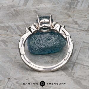 The Braided "Zuri" ring in 14k white gold with a 1.15-carat Montana sapphire is displayed upright on a rough blue stone, set on a textured gray surface with the Earth’s Treasury logo beneath.