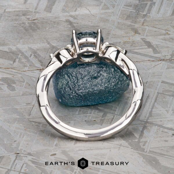 The Braided "Zuri" ring in 14k white gold with 1.15-carat Montana sapphire The Braided "Zuri" ring in 14k white gold with a 1.15-carat Montana sapphire is displayed upright on a rough blue stone, set on a textured gray surface with the Earth’s Treasury logo beneath.