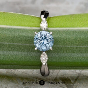 The Braided "Zuri" ring in 14k white gold features a 1.15-carat Montana sapphire center and two marquise-cut clear stones, beautifully displayed on a green leaf.