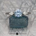 The "Zuri" ring in platinum, featuring a 0.87-carat Montana sapphire and two side stones, is showcased on rough blue crystal with a geometric metallic background. The image includes the text: "Earth's Treasury.