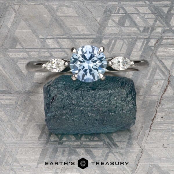 The "Zuri" ring in platinum with 0.87-carat Montana sapphire The "Zuri" ring in platinum, featuring a 0.87-carat Montana sapphire and two side stones, is showcased on rough blue crystal with a geometric metallic background. The image includes the text: "Earth's Treasury.