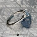 The "Zuri" ring in platinum, featuring a 0.87-carat Montana sapphire, rests on a rough blue stone atop a metallic patterned surface. The "Earth's Treasury" logo appears at the bottom.