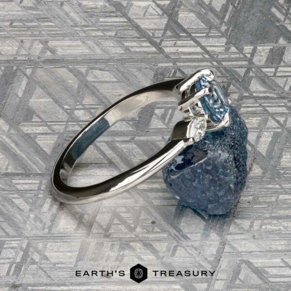 The "Zuri" ring in platinum with 0.87-carat Montana sapphire The "Zuri" ring in platinum, featuring a 0.87-carat Montana sapphire, rests on a rough blue stone atop a metallic patterned surface. The "Earth's Treasury" logo appears at the bottom.