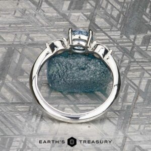 The "Zuri" ring in platinum with a 0.87-carat Montana sapphire is displayed upright on a textured gray surface, with the "Earth's Treasury" logo and text at the bottom.