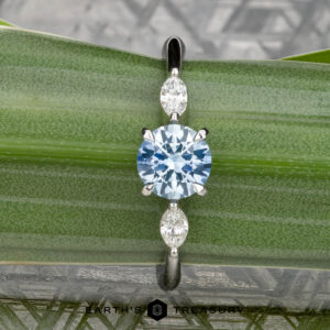 The "Zuri" ring in platinum features a 0.87-carat Montana sapphire center and two marquise-cut clear stones, beautifully displayed on a green leaf background.