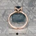 The "Musette" ring in 20k pink gold, featuring a 3.21-carat Montana sapphire, stands upright on a rough blue stone with a patterned gray backdrop. "Earth's Treasury" is displayed at the bottom.