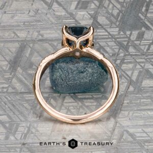 The "Musette" ring in 20k pink gold, featuring a 3.21-carat Montana sapphire, stands upright on a rough blue stone with a patterned gray backdrop. "Earth's Treasury" is displayed at the bottom.