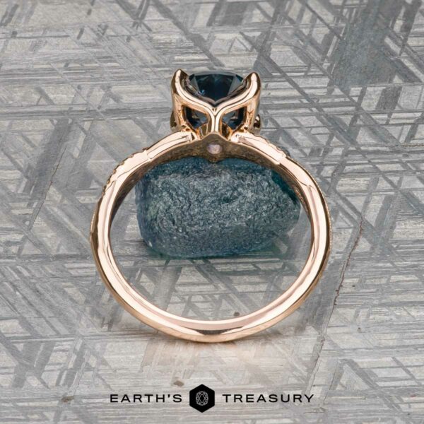 The "Musette" ring in 20k pink gold with 3.21-carat Montana sapphire The "Musette" ring in 20k pink gold, featuring a 3.21-carat Montana sapphire, stands upright on a rough blue stone with a patterned gray backdrop. "Earth's Treasury" is displayed at the bottom.