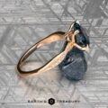 The "Musette" ring, crafted in 20k pink gold with a 3.21-carat Montana sapphire and accent diamonds, is shown on a textured metallic surface beside a rough blue stone, with The Earth’s Treasury logo at the bottom.