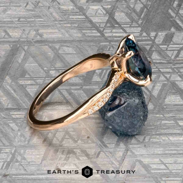 The "Musette" ring in 20k pink gold with 3.21-carat Montana sapphire The "Musette" ring, crafted in 20k pink gold with a 3.21-carat Montana sapphire and accent diamonds, is shown on a textured metallic surface beside a rough blue stone, with The Earth’s Treasury logo at the bottom.
