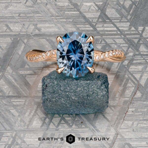 The "Musette" ring in 20k pink gold with 3.21-carat Montana sapphire The "Musette" ring in 20k pink gold with a 3.21-carat Montana sapphire rests on a rough stone atop a geometric surface. "EARTH'S TREASURY" is shown at the bottom.