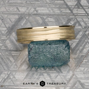 The "Silas" band in 14k yellow gold rests atop a rough blue gemstone on a patterned gray background, with the text "EARTH'S TREASURY" below.