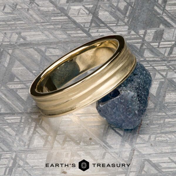 The "Silas" band in 14k yellow gold The "Silas" band in 14k yellow gold sits on a textured blue stone atop a metallic patterned surface, with the "Earth's Treasury" logo and text visible at the bottom.